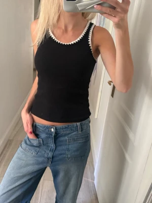 Svart ribbad linnetopp med vit kant  - Svart ribbad linnetopp med vit, taggig kant runt halsringningen. Toppen är ärmlös och har en tight passform som framhäver figuren. Perfekt att styla med jeans eller kjol för en trendig look.