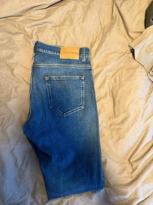 Blå raka jeans från Tiger of Sweden - Snygga blå jeans från Tiger of Sweden med klassisk femficksdesign och raka ben. Jeansen har en läderpatch med logga bak i midjan och subtila slitningar för en cool look. Perfekta till vardagsstilen och tillverkade i bomullsdenim.