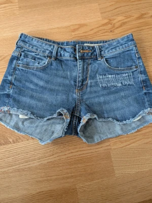 Blå jeansshorts med fransig kant - Snygga blå jeansshorts med fransig kant och slitningar framtill. Klassisk femficksmodell med knapp och dragkedja. Perfekta för sommaren och ger en avslappnad vibe. Passar till allt från t-shirt till hoodie.