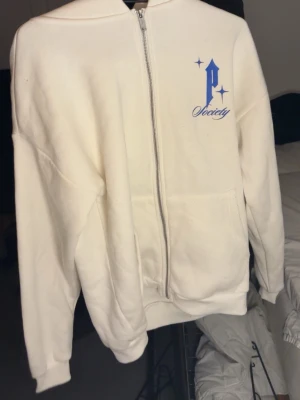 Vit zip-hoodie  - Vit hoodie med dragkedja från Paranoid Society. Tryck med blå text och duva på ryggen, samt blå logga på bröstet. Mjuk bomullskänsla, fickor fram och avslappnad passform. Perfekt för en streetwear-look.