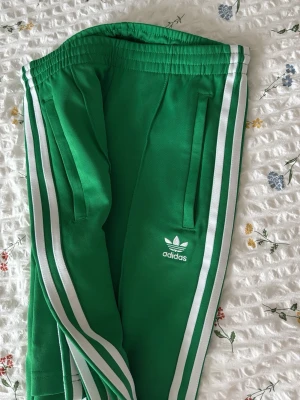 Gröna Adidas byxor med vita ränder - Snygga gröna träningsbyxor från Adidas med klassiska vita ränder längs sidorna och Adidas-logga på låret. Byxorna har resår i midjan och raka ben. Perfekta för sport eller chill. Materialet är troligtvis polyester för en sportig känsla.