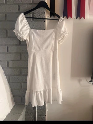 Vit kortklänning med puffärmar - Superfin vit kortklänning med puffärmar och volangkant nertill. Klänningen har fyrkantig urringning och snörning i ryggen för en extra snygg detalj. Perfekt för sommarens alla tillfällen! Klänningen är använd 1 gång på en skolavslutning 
