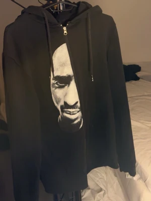 Svart zip-hoodie med tupac tryck - En svart hoodie med dragkedja och huva. Klassisk modell med långa ärmar och justerbara snören i huvan. Perfekt för en avslappnad stil och enkel att matcha med andra plagg.