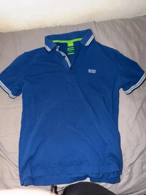 Hugo Boss piké  - Snygg blå pikétröja från BOSS med vit logga på bröstet. Tröjan har klassisk krage med knappar och vita/grå detaljer på krage och ärmslut. Perfekt för en stilren och avslappnad look.