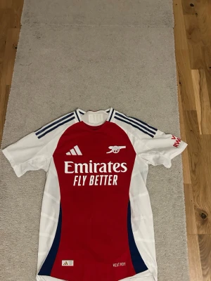 Arsenal matchtröja J. Timber 12 - Snygg Arsenal matchtröja från Adidas med J. Timber och nummer 12 på ryggen. Tröjan är röd med vita ärmar, blå detaljer och klassiska Adidas-ränder på axlarna. Tillverkad i funktionsmaterialet HEAT.RDY för bästa komfort under träning eller match.