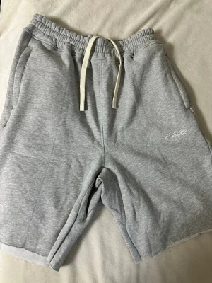 Grå sweatshorts från Corteiz - Grå sweatshorts från Corteiz med vit snörning i midjan och broderad logga framtill. Mjukt material med två sidofickor och en bakficka. Perfekta för chill dagar eller träning. Elastisk midja för skön passform. Säljer då de ej passar mig.