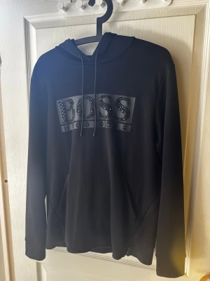 Svart hoodie från BOSS med nitar - Svart hoodie från BOSS med stort logotryck framtill dekorerat med coola nitar. Klassisk huva med dragsko och långa ärmar. Perfekt för en trendig och avslappnad stil ny pris:2600kr.