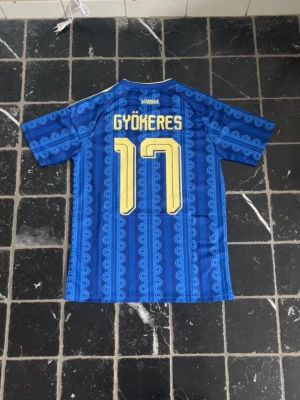 Blå Sverige fotbollströja Gyökeres 17 - Säljer en blå Sverige fotbollströja från Adidas med Gyökeres och nummer 17 i gult på ryggen. Tröjan har gula detaljer, klassiska Adidas-ränder på axlarna och Sveriges landslagsemblem på bröstet. Snyggt mönster i tyget och korta ärmar.
