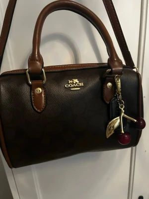 Brun handväska från Coach - Mörkbrun handväska från Coach i klassiskt läder med ljusbruna detaljer och guldfärgade metalldelar. Väskan har både handtag och axelrem samt ett körsbärsformat väskhänge. Perfekt för dig som gillar stilrena accessoarer.
