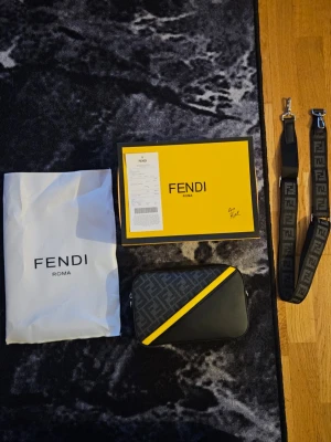 Fendi svart och gul handväska - Snygg handväska från Fendi i svart och grått med ikoniskt FF-mönster och en diagonal gul detalj. Väskan har en bred axelrem med Fendi-logga och levereras med originalpåse och ask. Perfekt för dig som gillar lyxiga accessoarer och vill sticka ut.