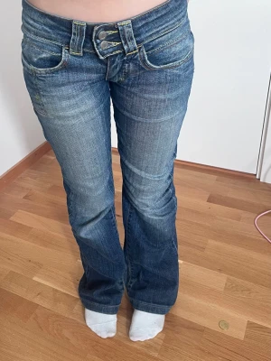 Blå bootcut jeans med dubbla knappar - Snygga blå bootcut jeans med dubbla knappar i midjan och klassiska fickor både fram och bak. Jeansen har en lätt tvättad look och markerade sömmar. Perfekta för dig som gillar en avslappnad men trendig stil.