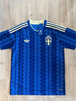 Sverige VM-2026 Fotbollströja - Säljer Sveriges bortatröja med Sveriges landslagsemblem på bröstet. Tröjan har gula ränder på axlarna, retroinspirerat mönster och korta ärmar. Materialet är lätt och andas, perfekt för träning eller match. Snygg gul detalj i kragen. HELT NY