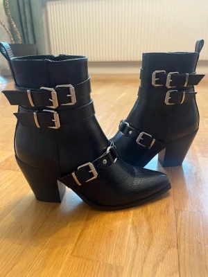 Svarta stövletter med spännen och klack - Snygga svarta stövletter med chunky klack och flera silvriga spännen. Skorna har spetsig tå och är gjorda i ett läderliknande material med struktur. Perfekta för dig som gillar en edgy stil och vill sticka ut lite extra. Storlek 39 i båda skorna nästan oanvända. Går att köpa bara ett par!