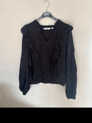 Svart volangblus från Kappahl  - Svart blus från Second Female med långa ärmar och volangdetaljer längs axlarna och framtill. V-ringad hals med knytband och lätt rynkad struktur. Perfekt för en trendig och stilren look.