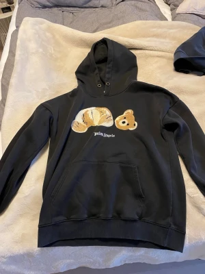 Svart hoodie Palm Angels med björn - Svart hoodie från Palm Angels med stor huva och känguruficka. På bröstet finns ett broderat motiv av en isärplockad nallebjörn och vit Palm Angels-logga under. Mjuk bomullskänsla och avslappnad passform, perfekt för en cool streetwear-look.