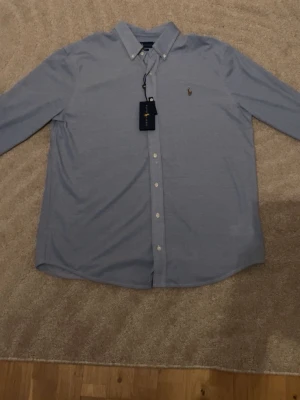 Blå skjorta från Polo Ralph Lauren - Klassisk ljusblå skjorta från Polo Ralph Lauren med button-down krage och broderad logga på bröstet. Skjortan har långa ärmar, vita knappar och är tillverkad i mjuk bomull. Perfekt för en clean och stilren look.