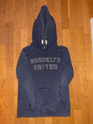 Schysst mörkblå hoodie med Brooklyn-tryck - Mörkblå hoodie med vit text 'BROOKLYN EQPT NYC' på bröstet. Klassisk modell med huva och känguruficka framtill. Mjuk bomullsblandning och ribbade muddar vid ärmslut och nederkant. Perfekt för en avslappnad streetstyle-look. Passar en kille som är 160-170 cm lång. 