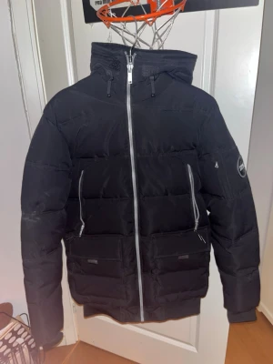 Svart pufferjacka från Supply & demand - LITE SMUTSIG PÅ BILD MEN RENGÖRS INNAN DEN SKICKAS!. Svart pufferjacka från Supply med hög krage, huva och silverfärgad dragkedja. Jackan har två stora fickor framtill och två snedställda bröstfickor med dragkedja. På vänster ärm finns en rund logotyp med texten 'SUPPLY'. Perfekt för kalla vinterdagar.