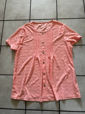 Korallfärgad kortärmad blus storlek XXL - Söt rosa blus med korta ärmar. Blusen har rund halsringning och knappar framtill. Lätt och luftig, perfekt för varma dagar. Materialet känns mjukt och har fina detaljer som ger en romantisk vibe.
