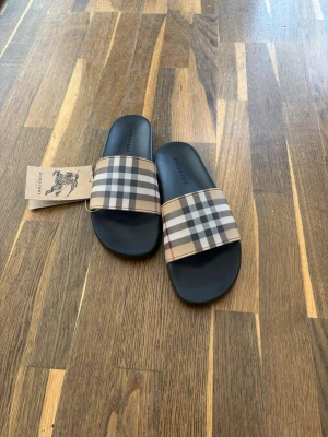 Burberry sandaler - Snygga sandaler från Burberry med klassiskt beige, svart och rött rutmönster på remmen. Svart sula i gummi och bred rem i textil med ikoniskt mönster. Perfekta för en trendig och avslappnad look.