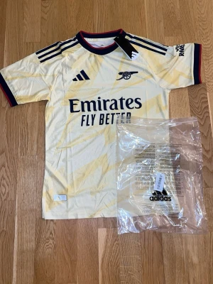Arsenal bortatröja Adidas gul - Snygg Arsenal bortatröja från Adidas i ljusgul med subtila mönster och mörkblå detaljer. Tröjan har rund hals med röd och blå kant, samt klubbmärke och Adidas-logga på bröstet. Tillverkad i lätt och ventilerande material, perfekt för träning eller match.