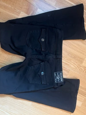 Svarta bootcut jeans True Religion - Säljer ett par svarta bootcut jeans från True Religion med klassiska fickor och silverfärgade knappar. Jeansen har utsvängda ben och tydliga sömmar, samt en patch med logga bak i midjan. Perfekta för dig som gillar en tidlös och cool stil. Byxorna är nästan helt nya, använda 1 gång därav prislappen som är borta. Säljer för dom va för korta för mig som är 178cm. Pris går att diskutera💕
