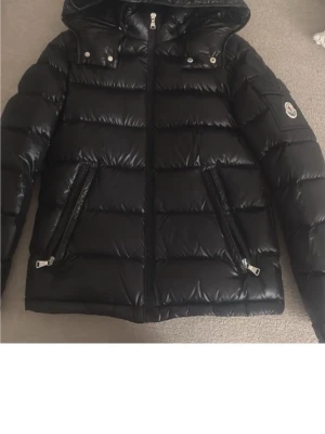 Svart dunjacka från Moncler - Snygg svart dunjacka från Moncler med glansig finish, huva och två dragkedjefickor framtill. Jackan har Moncler-logga på ärmen och klassisk quiltad design. Perfekt för kalla vinterdagar och riktigt stilren look.