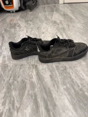 Svarta Nike Air Jordan 1 Low Travis Scott - Snygga svarta Nike Air Jordan 1 Low Travis Scott med vita kontrastsömmar och broderade detaljer på hälen. Skorna har klassisk swoosh-logga på sidan och är gjorda i mocka och läder. Perfekta sneakers för dig som gillar streetwear och unika samarbeten.