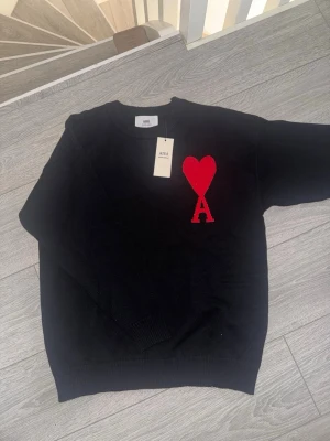 Svart stickad tröja från Ami paris - Svart stickad tröja från Ami Alexandre Mattiussi med röd hjärtlogga och bokstaven A på bröstet. Tröjan har rund halsringning och ribbade muddar. Perfekt för dig som gillar stilrena plagg med en twist.