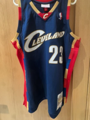 Cleveland Cavaliers basketlinne #23 Lebron James - Snyggt basketlinne från Mitchell & Ness Swingman Collection med Cleveland Cavaliers tryck och nummer 23. Marinblå bas med röda och gula detaljer, meshmaterial för bra ventilation. Perfekt för basketträning eller att bära som streetwear.