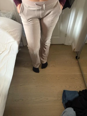 Beige velourbyxor från Juicy Couture - Säljer ett par beige mjukisbyxor i velour från Juicy Couture. Byxorna har utsvängda  ben, dragsko i midjan och fickor fram. På baksidan står det 'Juicy' i glittriga stenar. Perfekta för en chill och trendig look. På insidan har jag klippt ut på grund av att jag behövde fixa till dom men det är inget som märks passar både S och M 