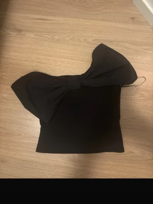 Svart topp med stor rosett från Monki - Supersnygg svart topp från Monki med en stor rosett på ena axeln. Toppen är axelbandslös och har en tight passform som ger en trendig look. Perfekt för dig som vill sticka ut med en unik detalj.