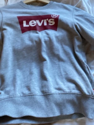Grå sweatshirt från Levi's (aldrig andvändt) - Snygg grå sweatshirt från Levi's med klassisk röd logga framtill. Tröjan har rund halsringning, långa ärmar och ribbade muddar vid ärmslut och nederkant. Perfekt för en avslappnad och trendig stil.