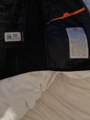 Svart dunväst från Parajumpers - Snygg svart dunväst från Parajumpers med quiltad design och orangea detaljer på insidan. Västen har hög krage och är lätt vadderad för extra värme. Perfekt för lager-på-lager och har märkets klassiska logga på insidan. Äkta !!