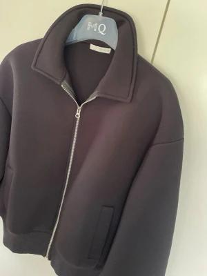Svart bomberjacka med dragkedja Zara  - Snygg svart bomberjacka med silverfärgad dragkedja och bred krage. Jackan har en minimalistisk design, ribbade muddar och raka fickor framtill. Perfekt för dig som gillar en clean och modern stil.