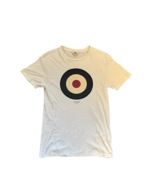 Ben Sherman T-shirt  - Vit t-shirt från Ben Sherman med ett stort grafiskt tryck i form av en måltavla på bröstet. Klassisk rund halsringning och korta ärmar. Tillverkad i mjuk bomull som känns skön mot huden. Perfekt för en avslappnad och stilren look.