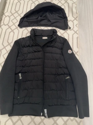 Svart pufferjacka från Moncler - Snygg svart pufferjacka från Moncler med avtagbar huva och ribbade ärmar. Jackan har två dragkedjeförsedda fickor framtill och Moncler-logga på vänster ärm. Materialet är en mix av quiltad nylon och stickad bomull för extra komfort och stil.