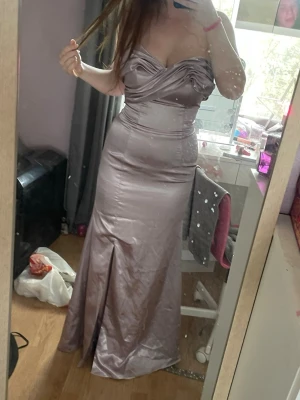 Lång lavendelfärgad satinklänning  - Säljer en elegant lång klänning i glansigt lavendelfärgat satinmaterial. Klänningen har en axelbandslös design med draperad byst och markerad midja. Kjolen är figurnära med en slits framtill för extra rörelsefrihet. Ungefär korset liknande precis över midjan. Den är även formad vid bysten. Perfekt för bal eller festkväll.