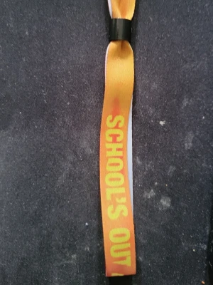 Festivalarmband School's Out VIP - Säljer ett festivalarmband i tyg med texten 'SCHOOL'S OUT' på ena sidan och 'VIP' på andra. Armbandet är i orange och rött med gula och vita bokstäver samt svart plastspänne. Perfekt som minne eller accessoar till festivaloutfiten.