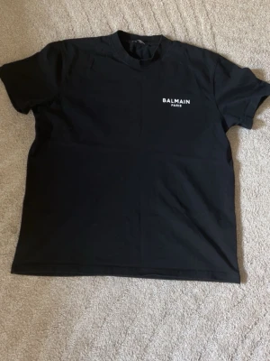 Svart Balmain Paris t-shirt - Snygg svart t-shirt från Balmain Paris med vit logga tryckt på bröstet. Klassisk passform och rund halsringning, perfekt för en stilren och trendig look. Tillverkad i mjuk bomull som känns skön mot huden.