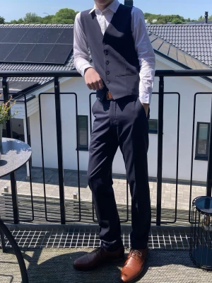 Mörkblå kostymväst och byxor - Snygg mörkblå kostymväst och matchande byxor, perfekt för dig som vill ha en stilren och klassisk look. Perfekt för student/skolavslutning. Västen har knappar framtill och byxorna har en rak passform. Bärs med vit skjorta för en clean outfit. Samt snygga finskor från Jack & Jones i cognacs-färg i storlek 44. Slimfit-byxor och väst från Selected i storlek 44. Skjorta från Dressmann i storlek XS. Hela outfiten har endast använts en gång. 