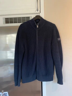 Lexington Company  Zip Up Sweater - Mörkblå stickad tröja från Lexington med dragkedja hela vägen och ståkrage. Tröjan har ribbstickad struktur och en patch med logga på ärmen. Perfekt för lager-på-lager och snygg till jeans.