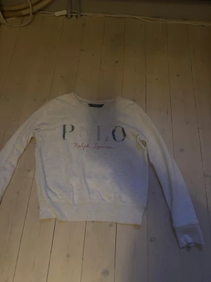 Grå sweatshirt från Polo Ralph Lauren - Ljusgrå sweatshirt med rund halsringning och tryckt POLO-logo framtill i blått och rött. Tröjan har långa ärmar och ribbade muddar vid ärmslut och nederkant. Perfekt för en avslappnad och stilren look.