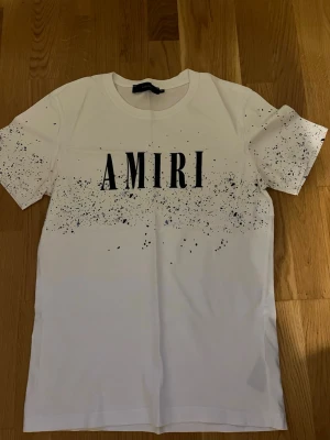 Vit AMIRI t-shirt med stänkmönster - Snygg vit t-shirt från AMIRI med svart logga framtill och coolt stänkmönster över bröstet och ärmarna. Klassisk passform med rund hals och korta ärmar. Perfekt för dig som gillar streetwear och vill sticka ut.