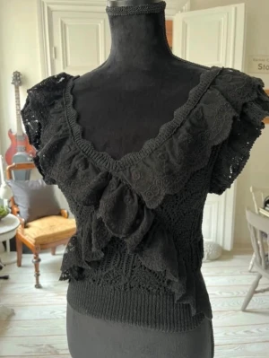 Svart virkad blus med volang - Supersöt svart virkad blus med v-ringning och volangdetaljer framtill och vid ärmarna. Kortärmad modell med romantisk känsla och ribbad nederkant. Perfekt för dig som gillar unika toppar med mycket detaljer.