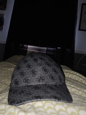 Grå keps med mönster från Guess - Snygg grå keps från Guess med all-over logomönster i svart. Klassisk böjd skärm och justerbar baksida. Perfekt accessoar för en trendig streetstyle-look.
