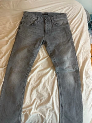 Grå Nudie Jeans grim Tim - Storlek 32/34. Säljer ett par grå jeans från Nudie Jeans med klassisk femficksmodell och raka ben. Jeansen har Nudie-loggan på bakfickan. Perfekta för dig som gillar en stilren och avslappnad look. Knappt använd någonting