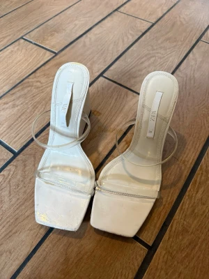 Vita klackar från Zara - Säljer ett par trendiga vita wedge-sandaler från Zara med transparenta remmar och fyrkantig tå. Skorna har hög kilklack och är tillverkade i syntetmaterial. Använda 1-2 gånger. Har fått små repor på sulan som man ser på bilden. Storlek 39.