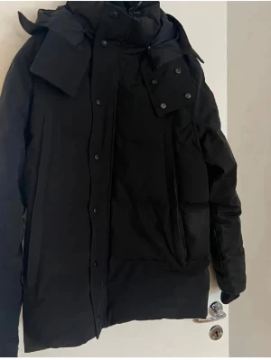 Canada goose wyndham  - Snygg svart dunjacka från Canada Goose med klassisk patch på ärmen. Jackan har hög krage, huva och flera praktiska fickor framtill. Perfekt för kalla vinterdagar och har en stilren, rak passform.