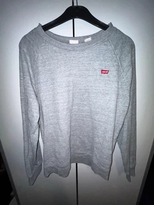 Grå sweatshirt från Levi's - En stilren grå sweatshirt från Levi's med klassisk rund halsringning och diskret röd logotyp på bröstet. Tröjan har långa ärmar och är tillverkad i mjukt material som passar perfekt till en avslappnad look.